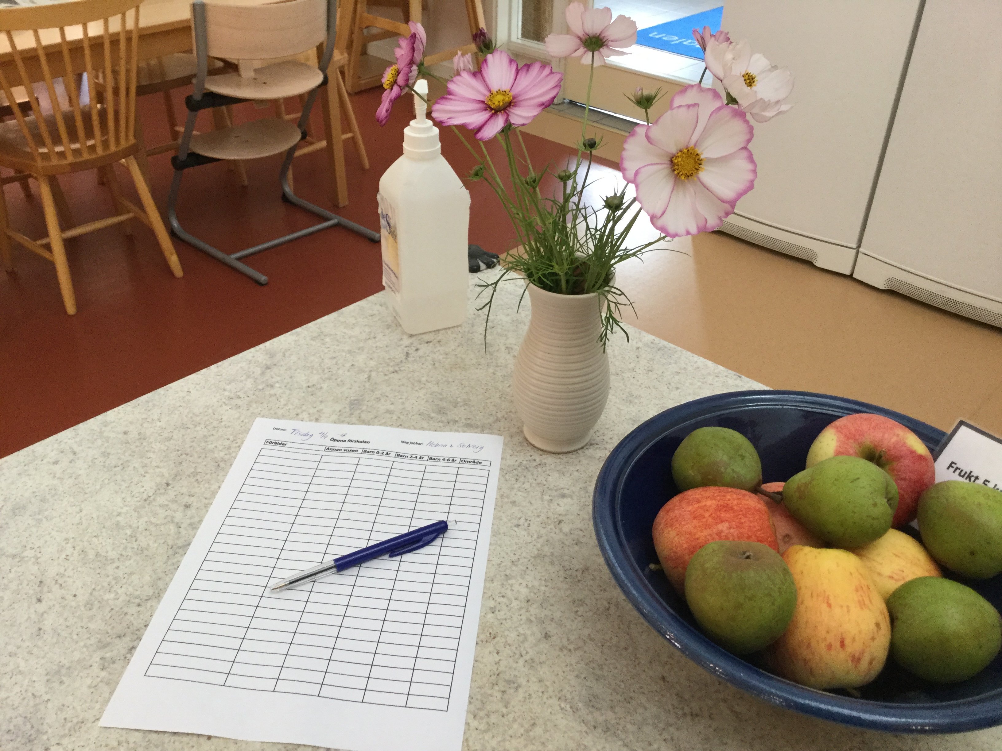 En bänk med närbild på en vas med blomma i, anmälningslista och ett fruktfat