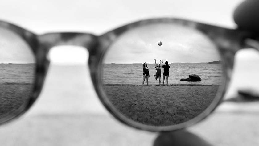 Svartvit bild på solglasögon i närbild, i bakgrunden tre personer på en strand som kastar boll