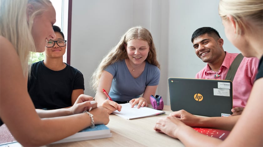5 glada ungdomar i samtal vid ett bord, dator och penna och papper på bordet 