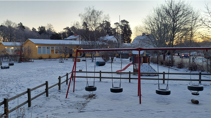 Översikt över gård och förskola i snötäcke, gungor längst fram i bilden