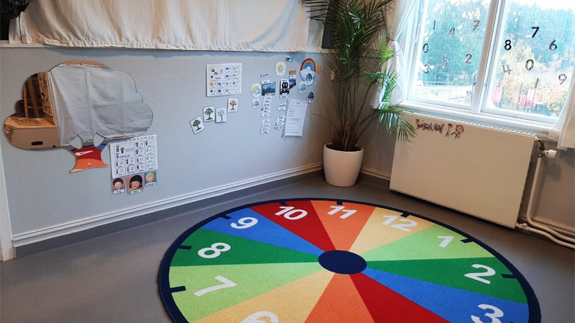 Bild på ett rum på förskolan, en matta i olika färger som en klocka och siffror på fönstren
