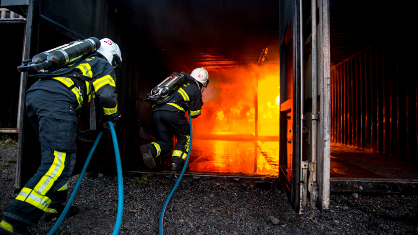 Två brandmän med slang på väg in i en brinnande container