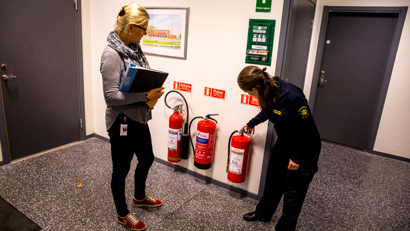 En kvinna inspekterar brandsläckare och en annan kvinna tittar på med en pärm i handen